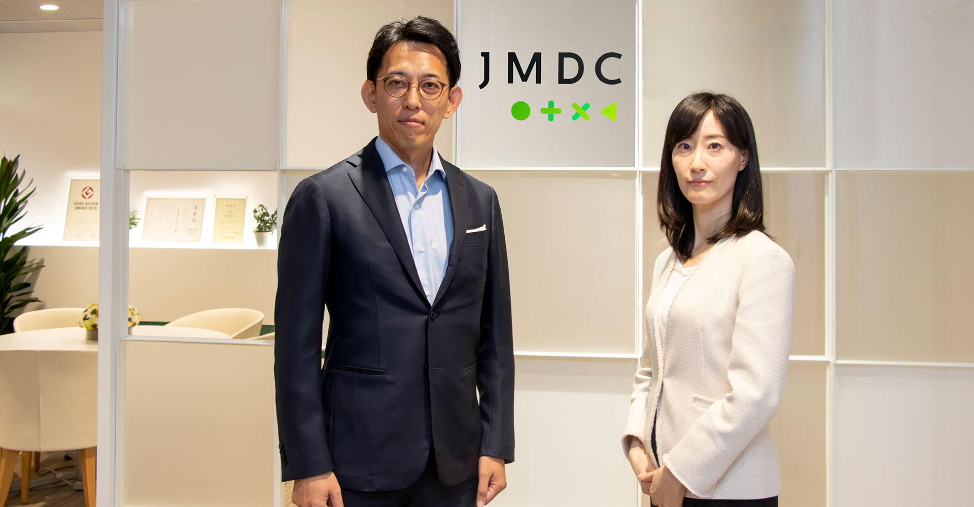 株式会社JMDCのロゴの前に立つ代表取締役 野口氏と聞き手、野村アセットマネジメント株式会社 鳥居